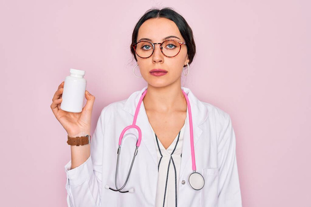Young beautiful doctor woman with blue eyes wearing stethoscope holding medicine pills with a confident expression on smart face thinking serious - 写真・画像