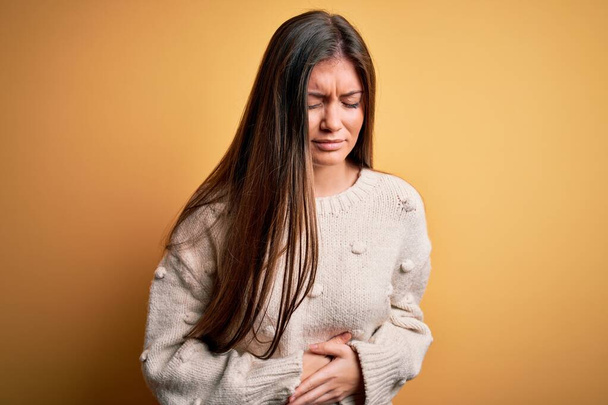 Young beautiful woman with blue eyes wearing casual sweater standing over yellow background with hand on stomach because indigestion, painful illness feeling unwell. Ache concept. - Foto, Bild