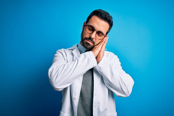 Young handsome doctor man with beard wearing coat and glasses over blue background sleeping tired dreaming and posing with hands together while smiling with closed eyes. - Foto, imagen