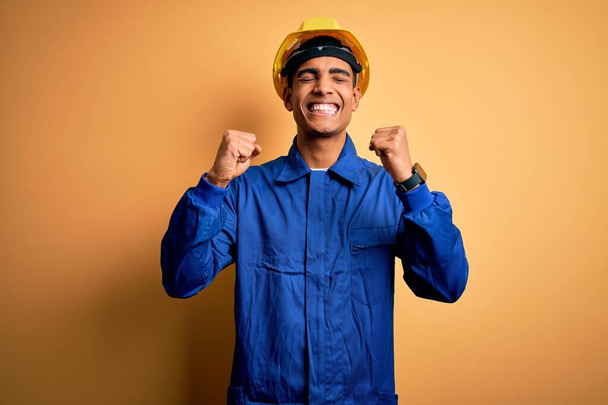 Νεαρός όμορφος Αφροαμερικάνος εργάτης με μπλε στολή και κράνος, ενθουσιασμένος για την επιτυχία με τα χέρια σηκωμένα και τα μάτια κλειστά, γιορτάζοντας τη νίκη χαμογελώντας. Έννοια νικητή. - Φωτογραφία, εικόνα