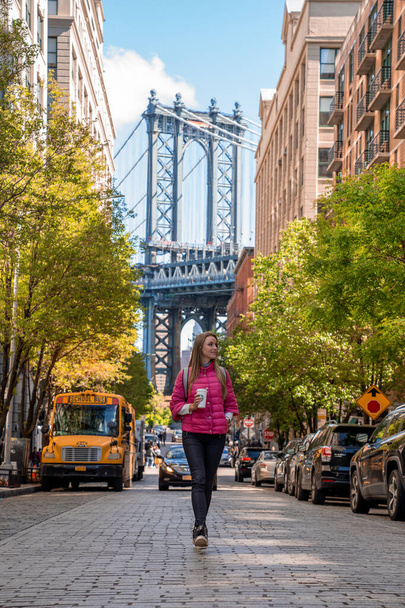 New York, USA. May 10, 2019.  Young girl walking down the street of Brooklyn drinking coffee with a Manhattan bridge in the background. - Фото, зображення