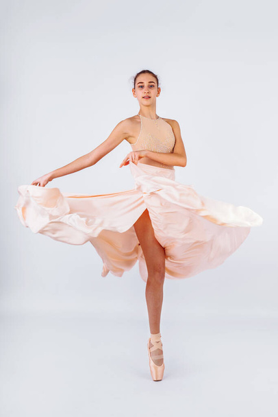Μια νεαρή και απίστευτα όμορφη μπαλαρίνα ποζάρει και χορεύει σε ένα λευκό στούντιο γεμάτο φως. Η ομορφιά της κλασικής τέχνης του μπαλέτου. Χορός με χορευτική μπαλαρίνα, ποζάρει σε τρυφερό tutu ροδάκινο - Φωτογραφία, εικόνα