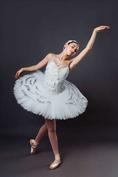 Portrait of young beautiful graceful caucasian ballerina practice ballet positions in tutu skirt of white swan from Swan Lake. Classical Ballet dancer in studio on gray background standing on tiptoe - Photo, Image