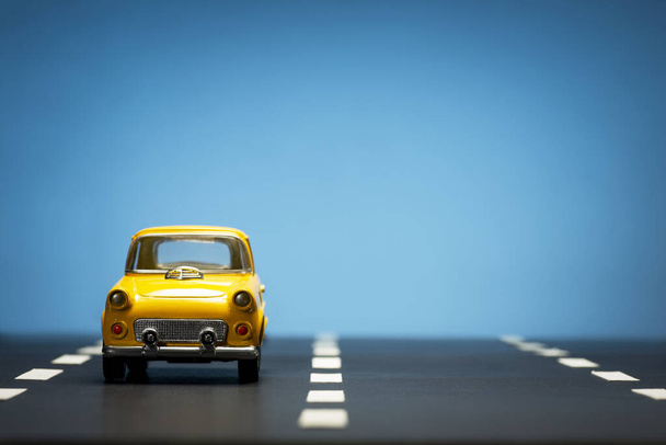 Frontansicht eines gelben Spielzeugautos auf blauem Hintergrund und auf einer asphaltierten Straße. - Foto, Bild
