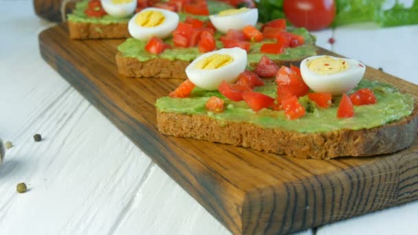 Healthy Vegan food. Spread mashed avocado on toasted brown bread sprinkled with black and red pepper. Making tasty avocado toast for breakfast. Cooking bruschetta with cherry tomatoes and quail eggs. - Footage, Video