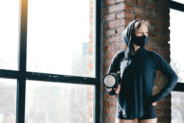 Trainer in black hoodie and mask holds a black massage cylinder and looks through picture window. Rest during a workout at home. - Valokuva, kuva