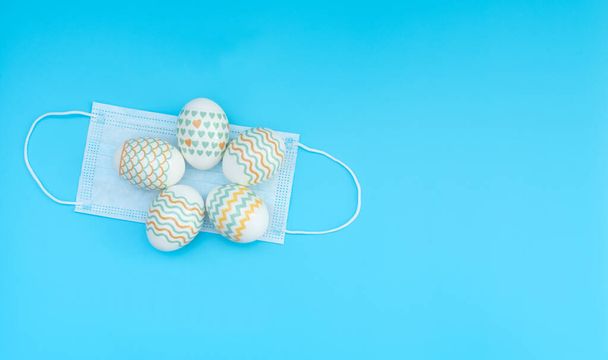 Decorated Easter eggs lie on the medical face mask on blue background with copy space. Happy Easter during quarantine concept. Biohazard, 2019-nCoV, COVID-19, social distance, stay home. Flat lay. - Foto, Imagen