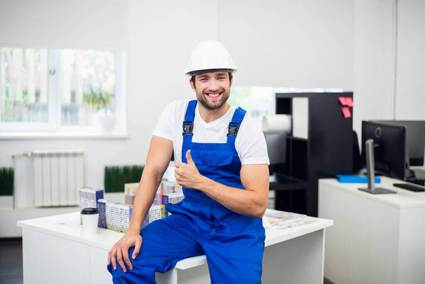 Başparmaklarını gösteren sert şapkalı gülümseyen bir inşaat işçisinin portresi. - Fotoğraf, Görsel
