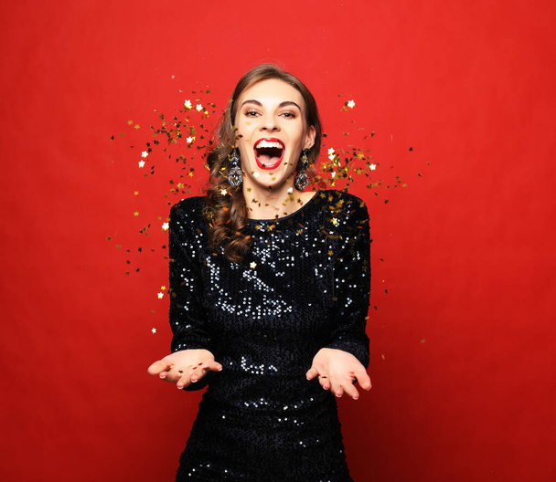 Holiday concept: beautiful brunette female wearing evening dress standing under confetti rain over red background - Fotó, kép