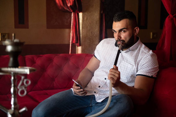 Young Man Smoking Shisha At Arabic Restaurant - Man Exhaling Smoke Inhaling From A Hookah and Texting on Phone - Photo, image