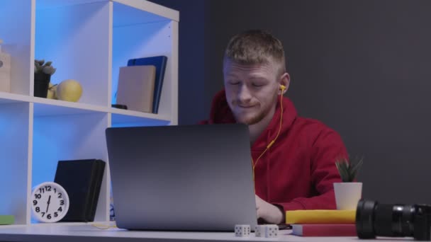 Unavený atraktivní muž na volné noze se sluchátky pracuje na notebooku pozdě v noci - Záběry, video