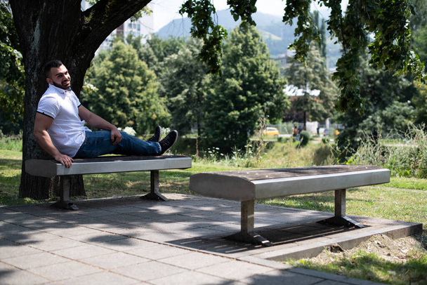 Porträt eines selbstbewussten und erfolgreichen jungen Mannes mit weißem Hemd im Park, der auf einer Bank sitzt - Foto, Bild