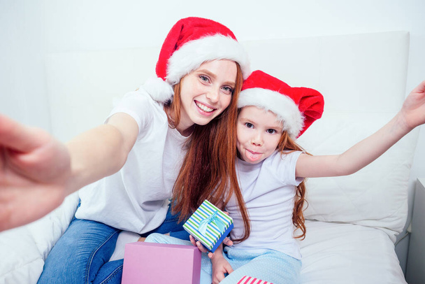 beautiful redhaired ginger woman in santa claus hat and happy little girl in pajamas sitting on bed with gifts boxes,taking selfie pictures on smartphone camera.christmas morning celebration - Foto, afbeelding