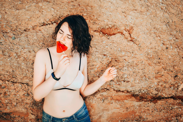 A young sexy girl licks a caramel candy on a heart shaped stick while standing against a stone background. - Фото, зображення