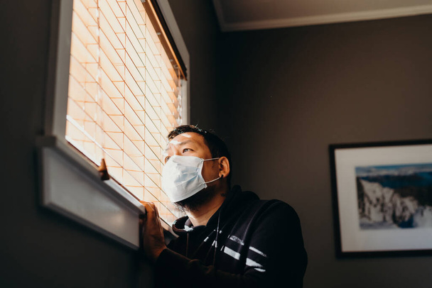 Азіат на самоті, дивлячись через вікна на жалюзі. Людина в медичній масці змушена залишатися вдома в результаті обмежень, спричинених спалахом коронавірусу.. - Фото, зображення