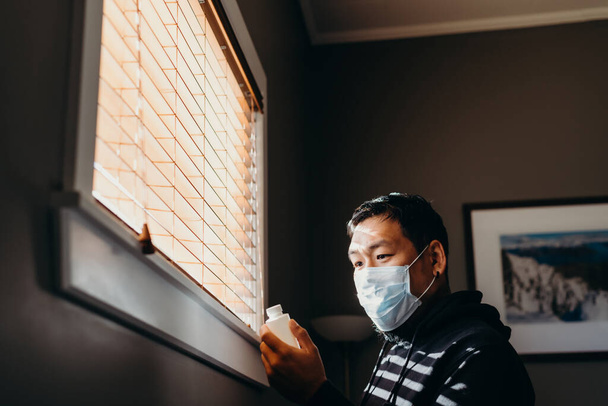 Homem asiático em isolamento olhando para garrafa de remédio. Homem de máscara médica forçado a ficar dentro de casa como resultado das restrições causadas pelo surto de Coronavirus
. - Foto, Imagem