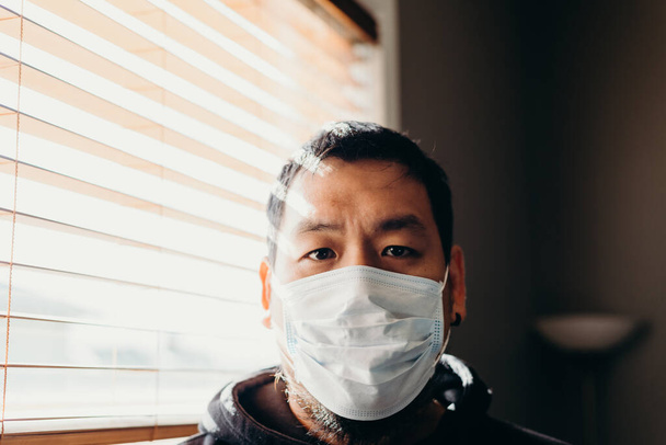 Asiatique dans l'isolement regardant la caméra et se sentant triste. Homme sous masque médical forcé de rester à l'intérieur de la maison en raison des restrictions causées par l'épidémie de coronavirus
. - Photo, image
