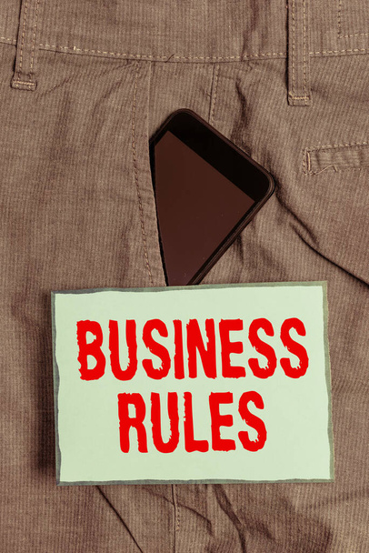 Χειρόγραφο κείμενο που γράφει τους κανόνες των επιχειρήσεων. Έννοια σημαίνει μια συγκεκριμένη οδηγία που περιορίζει ή ορίζει μια συσκευή Smartphone επιχείρησης μέσα σε επίσημο παντελόνι εργασίας μπροστινή τσέπη κοντά σε χαρτί σημείωμα. - Φωτογραφία, εικόνα