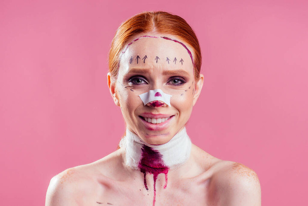 medical insurance concept. happy redhaired ginger woman with lines on her face and bandage blood on neck in studio pink background - Foto, Imagem