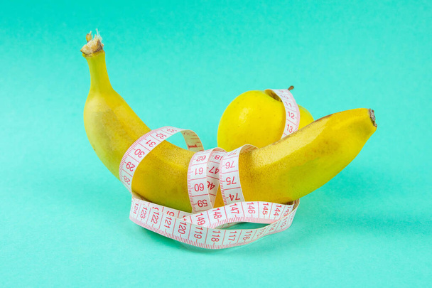 Verschiedene Obstsorten wie Äpfel und Bananen liegen auf einem einfüßigen Sockel und sind mit einem Maßband umwickelt - ein Konzept für eine gesunde Gewichtsabnahme, das Spaß macht. - Foto, Bild