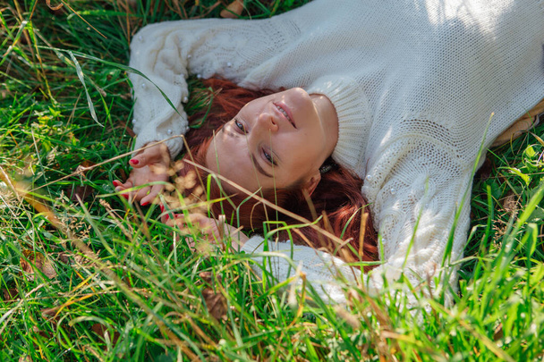 Morning portrait of a beautiful happy woman with red hair laying in grass in white sweater - Foto, Imagem