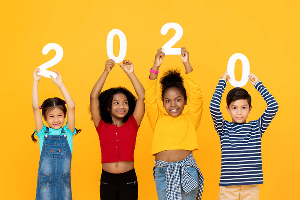 Yeni yıl konsepti için gülümseyen ve 2020 rakamı tutan sevimli melez çocuklar sarı arka planda izole edilmiş. - Fotoğraf, Görsel