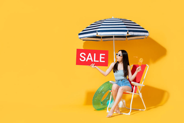 Здивована красива азіатка влітку, тримаючи червоний знак продажу, сидячи на пляжному стільці ізольованому на жовтому тлі з копіювальним простором. - Фото, зображення
