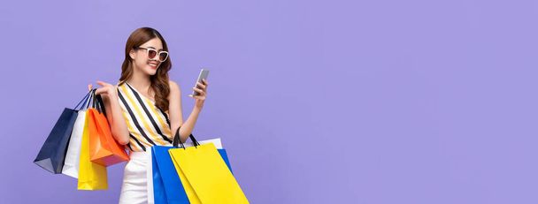 Moda hermosa mujer asiática joven llevando coloridas bolsas de compras en línea con el teléfono móvil aislado en el fondo de la bandera púrpura
 - Foto, Imagen