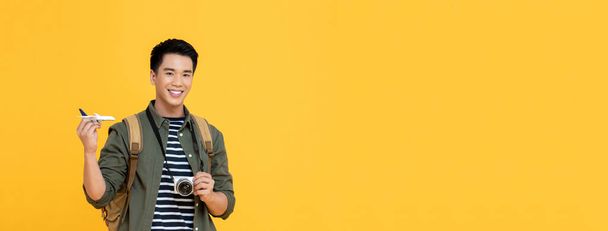 Young handsome smiling Asian tourist man holding plane model and camera isolated on yellow banner background with copy space - Фото, изображение