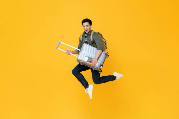 Joven turista asiático emocionado con el equipaje saltando en el aire listo para viajar aislado sobre fondo amarillo
 - Foto, imagen