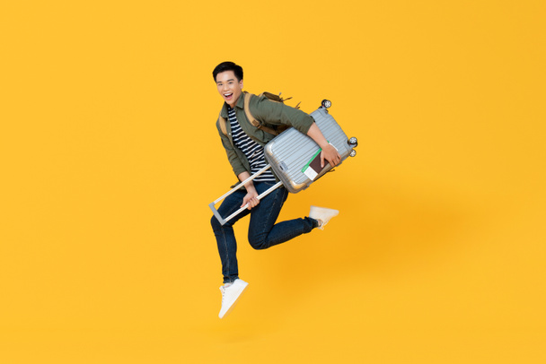 młody podekscytowany azjatycki turysta z bagażu skacze w powietrzu gotowy do podróży odizolowany na żółtym tle - Zdjęcie, obraz