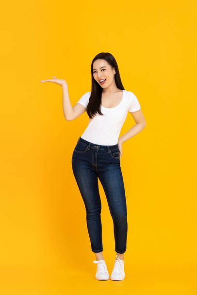 Retrato de comprimento total de sorrir confiante jovem bonita modelo feminino asiático fazendo um gesto de palma aberta em um fundo estúdio isolado amarelo
 - Foto, Imagem