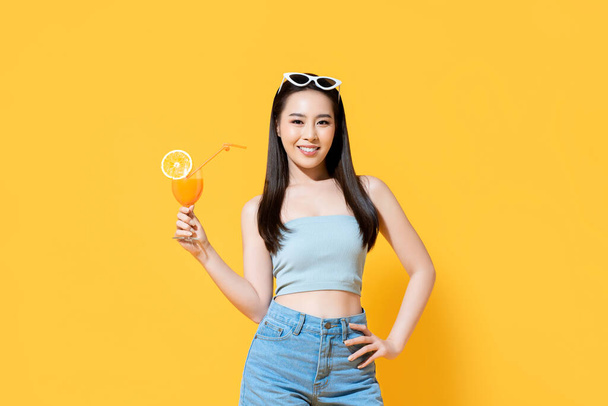 Schöne lächelnde Asiatin im Sommer-Outfit mit einem Glas Orangensaft auf farbenfrohem gelben Hintergrund - Foto, Bild