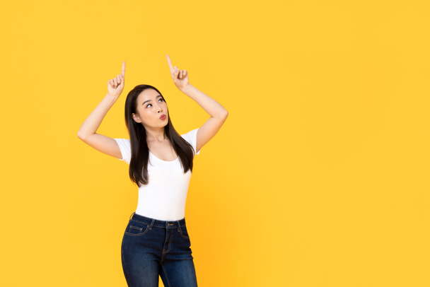 Jovem bela mulher asiática apontando para cima com seus dois dedos enquanto puxa os lábios em um fundo de estúdio isolado amarelo
 - Foto, Imagem