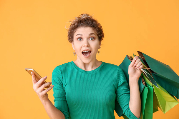 Surprised young woman with mobile phone and shopping bags on color background - Фото, зображення