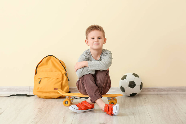 Cute little boy with skateboard, backpack and soccer ball sitting near color wall - Foto, afbeelding