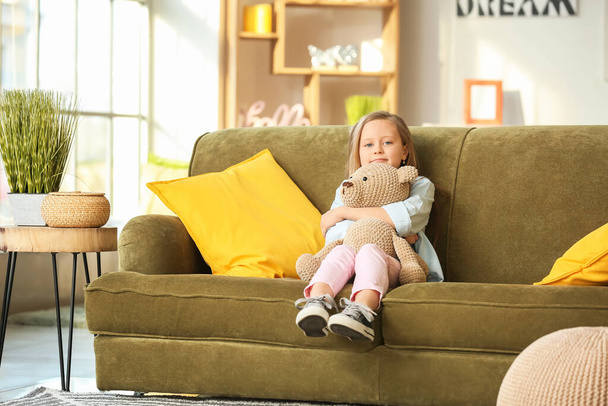 Cute little girl with teddy bear sitting on sofa at home - Фото, изображение