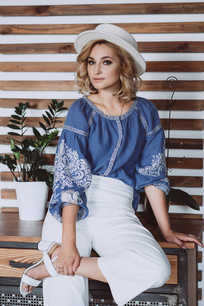 Mooi stijlvol blond meisje in etnische kleding in een geborduurd shirt met een witte muts in het interieur in grunge stijl. Oekraïense nationale kleding. - Foto, afbeelding