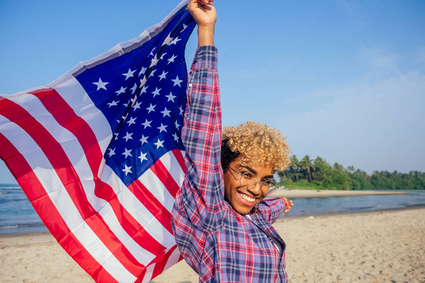 happy afro american woman holding usa flag at sunny tropical beach.4th of July Independence Day concept - Zdjęcie, obraz