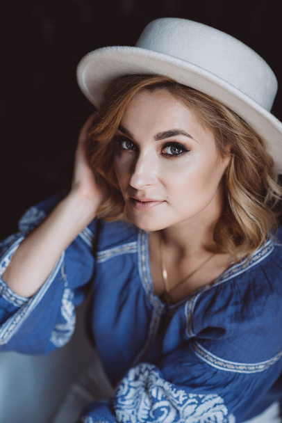 Beautiful stylish elegant blonde girl in ethnic clothes in an embroidered shirt in a white hat in the interior in grunge style. Ukrainian national clothes. - 写真・画像