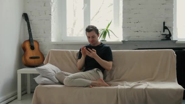 Man looking to smartphone sitting on sofa at home - Кадри, відео