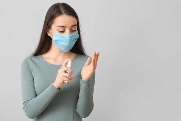Young woman wearing medical mask and using sanitizer against grey background - Photo, Image