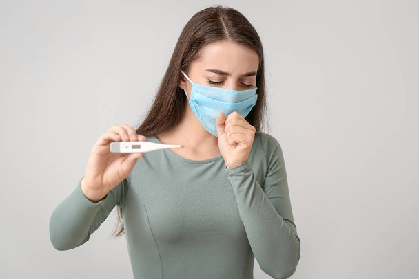 Ill young woman with medical mask and thermometer against grey background - Photo, Image