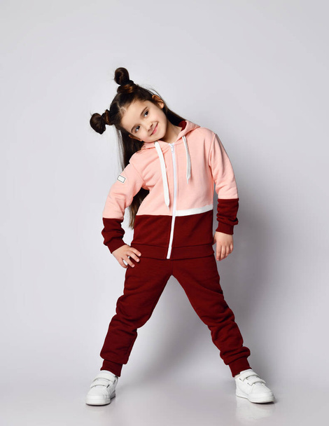 Sly smiling brunette kid girl with buns in modern stylish pink brown sport suit is posing with her head bowed aside - Fotó, kép