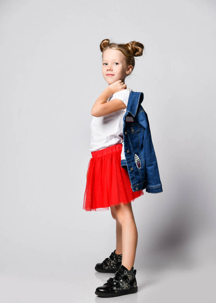 Smiling blonde kid girl with funny buns in red skirt and white t-shirt poses side to us with denim jacket over shoulder - Fotó, kép
