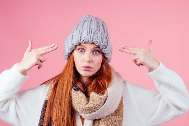 redhaired ginger woman in hat,knitted sweater and scarf pointing fingers hands like guns gesture at head in studio pink background copyspase - Photo, Image