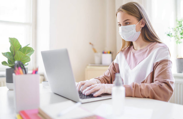 Young female student with face mask at the table, using laptop in quarantine. - Фото, изображение