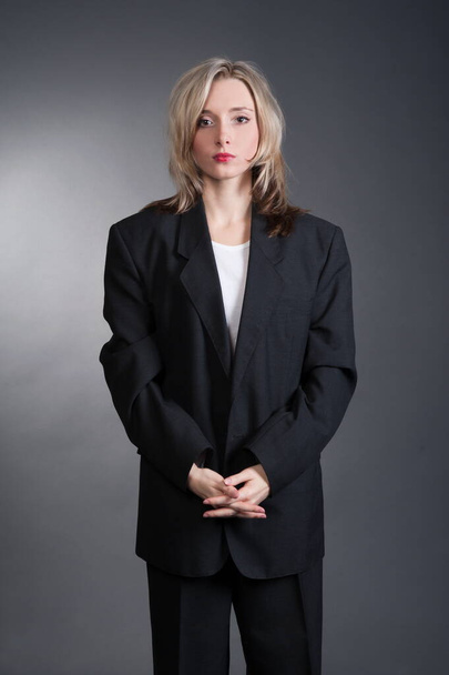 Schöne sexy junge blonde Frau in einem dunklen Business-Anzug, weißem Hemd und Krawatte posiert auf grauem Hintergrund - Foto, Bild