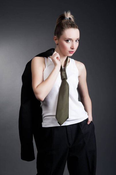 Beautiful sexy young blonde woman in a dark business suit, white shirt and tie posing on a gray background - Фото, изображение