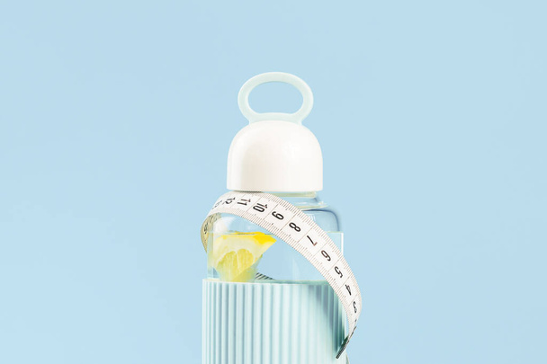 bílá páska měří kolem skleněné láhve s citronovou vodou a červené jablko na modré. Prostor pro kopírování předního pohledu. Zdravý životní styl, hubnutí a řízení a hydratace nebo dehydratace koncepce - Fotografie, Obrázek